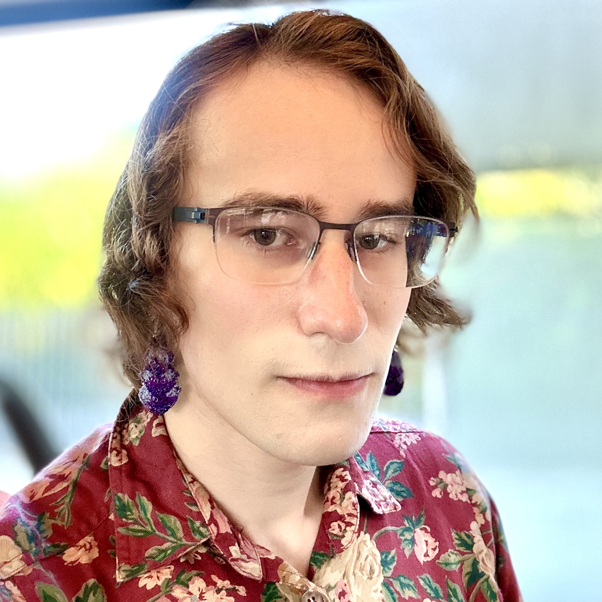 Portrait of a person (Nikolay Malkin in Singapore in 2025) with medium-length wavy brown hair, a floral shirt, purple earrings, and glasses smiling against a blurry green background.