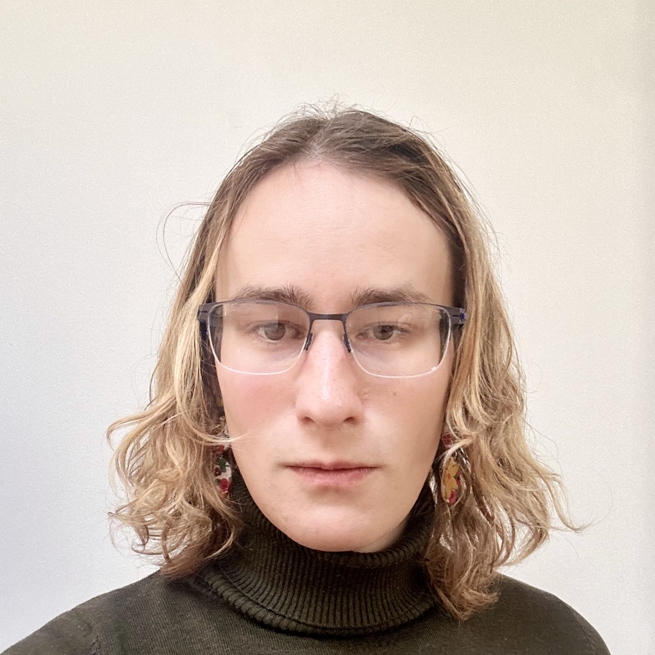 Portrait of a person (Nikolay Malkin in Montreal in 2025) with medium-length wavy brown hair, a green turtleneck, earrings, and glasses smiling against a white background.