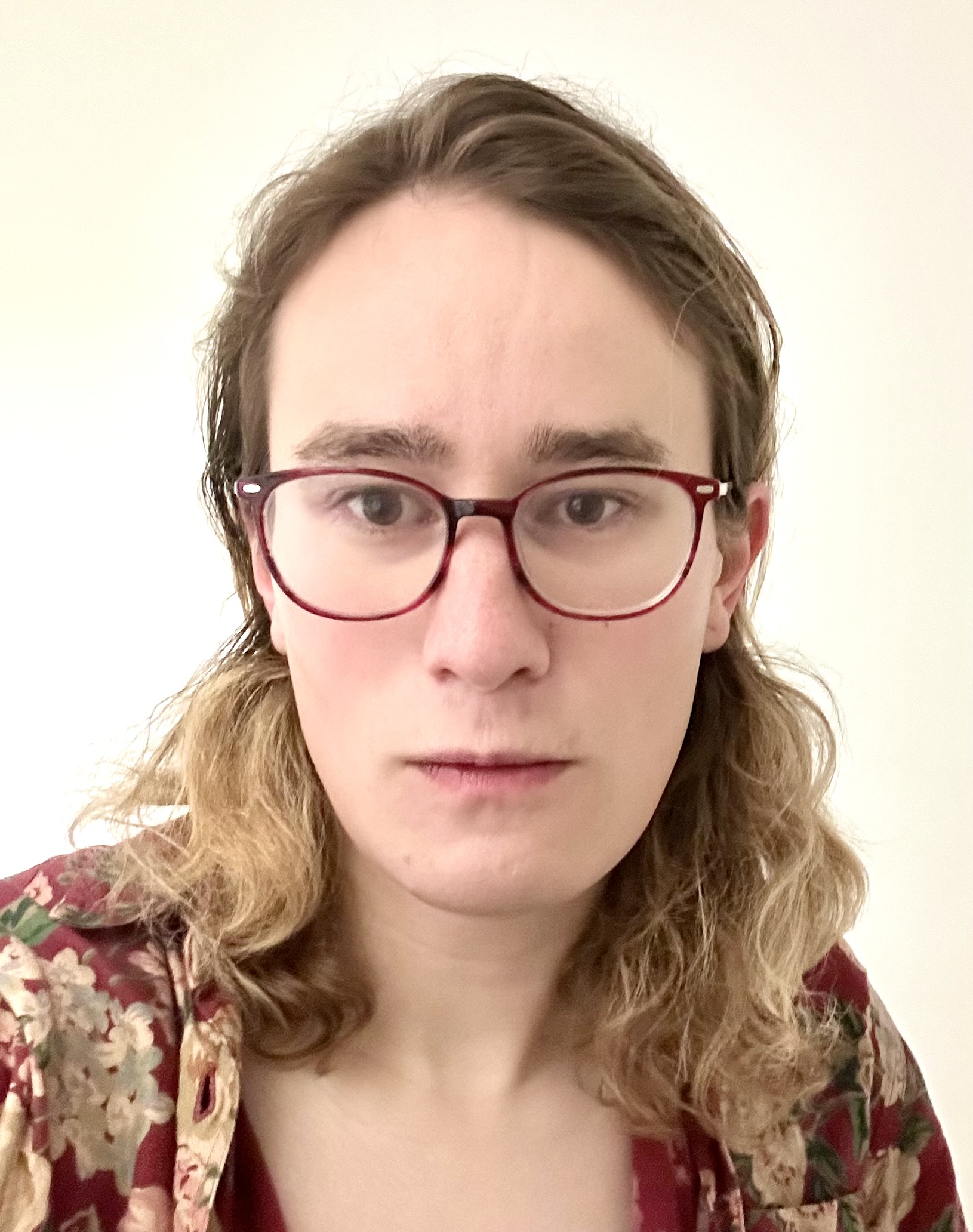 Portrait of a person (Esmeralda S. Whitammer, formerly called Nikolay Malkin, in early 2026) with shoulder-length wavy brown hair and glasses wearing a floral-patterned top against a plain light background.